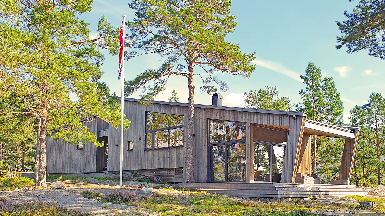 Nordvest fasade av hytte på Tjodholmen, med berg i forgrunn, trær tett opp mot hytten og flagg som vifter foran.